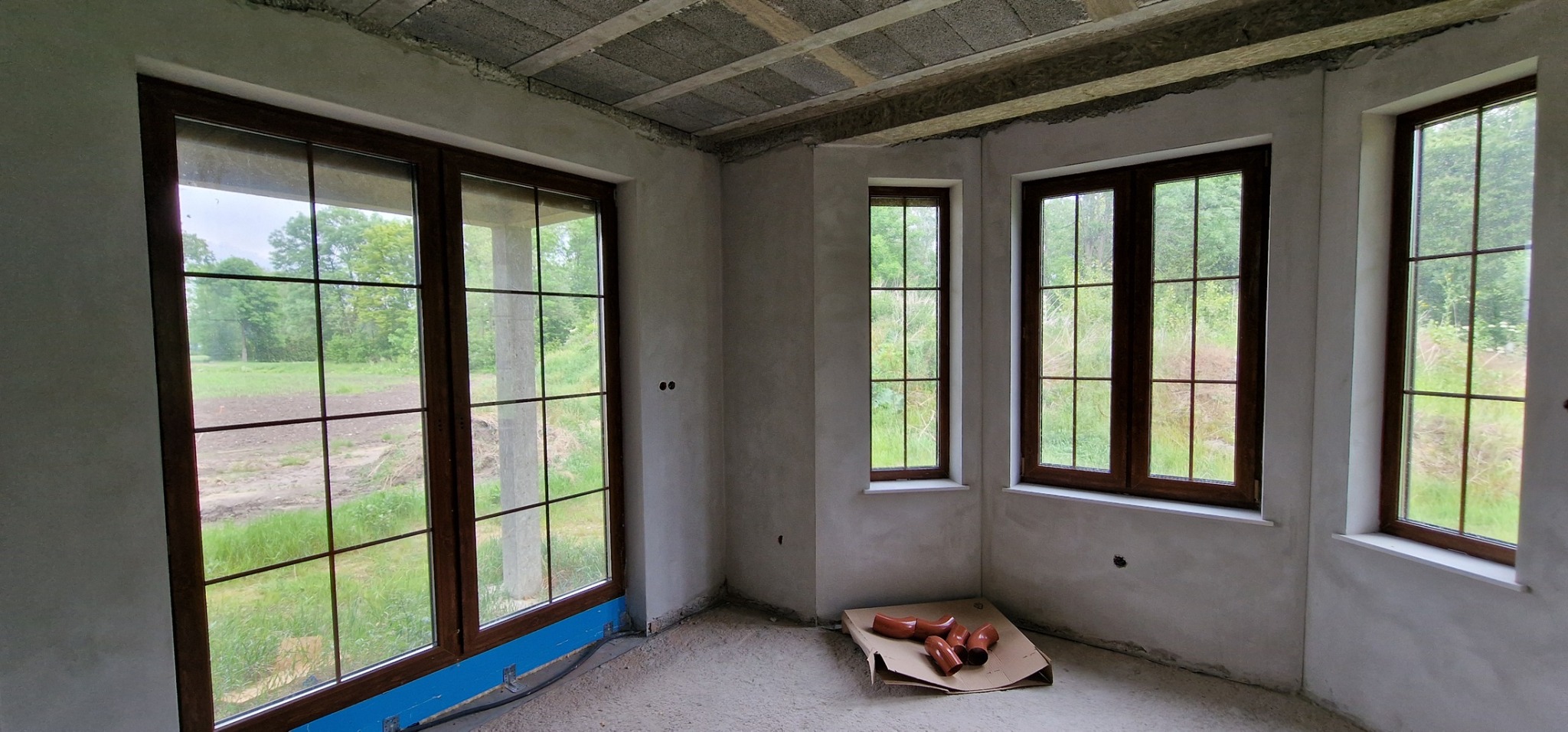 Wnętrze w stanie deweloperskim z trzema oknami PCV w kolorze ciemnego drewna, widok na zewnątrz na zieleń, na podłodze karton z rurami kanalizacyjnymi.