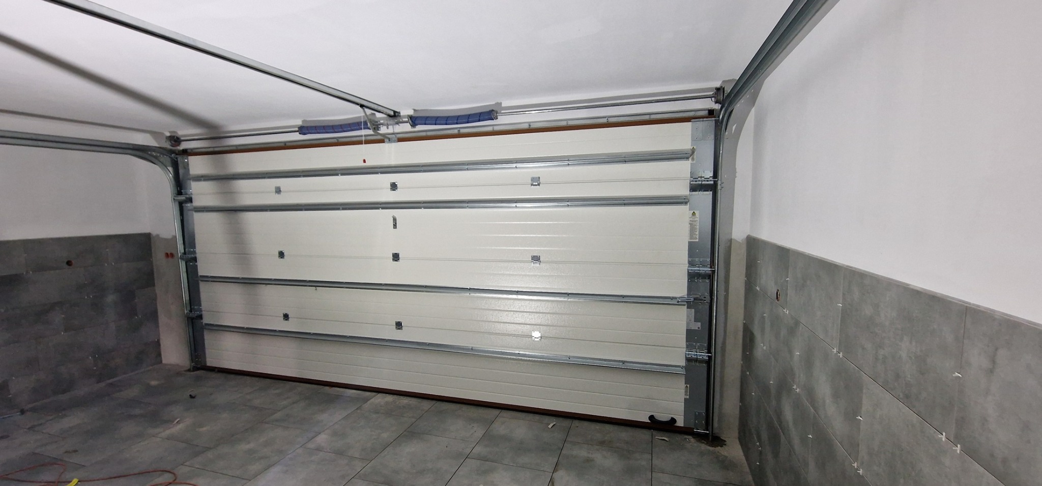 Zamknięta segmentowa brama garażowa w jasnym garażu z szarymi płytkami na podłodze i ścianach.