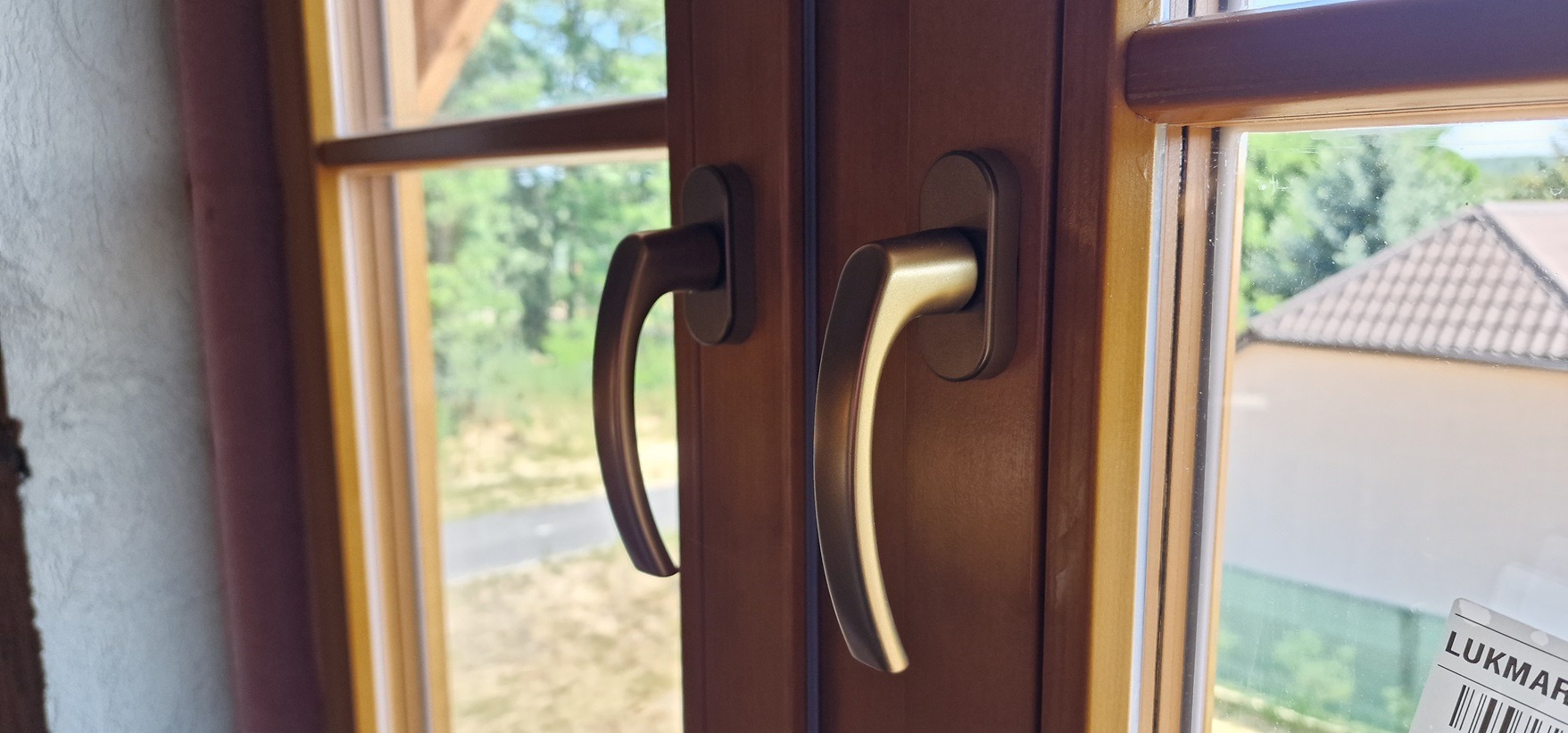 Brązowe okno PCV z dwiema klamkami, widok na zewnątrz z zielenią i budynkiem w tle, naklejka producenta w rogu szyby.