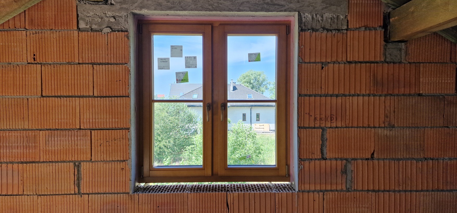 Montaż drewnianego okna dwuskrzydłowego w surowym murze z cegły ceramicznej; widoczne naklejki producenta na szybie i fragmenty zaprawy murarskiej.