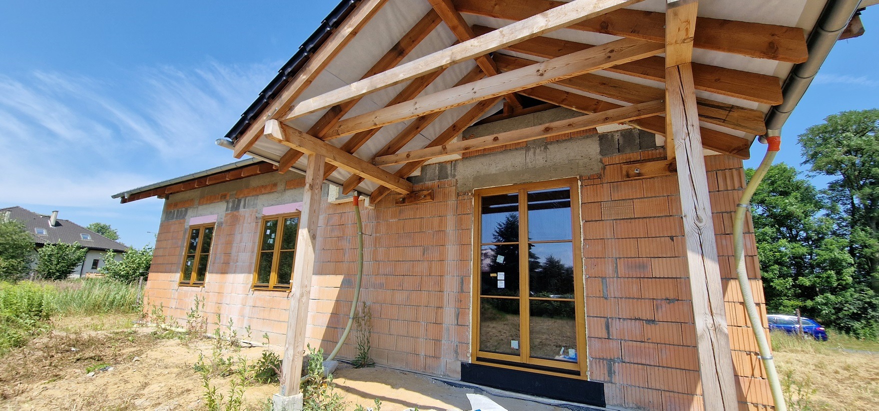 Surowy stan budynku w trakcie budowy z widocznymi drewnianymi oknami i konstrukcją dachu, ceglana elewacja, nieotynkowane ściany, rynna z pomarańczową taśmą. Widoczne elementy konstrukcyjne i brak...