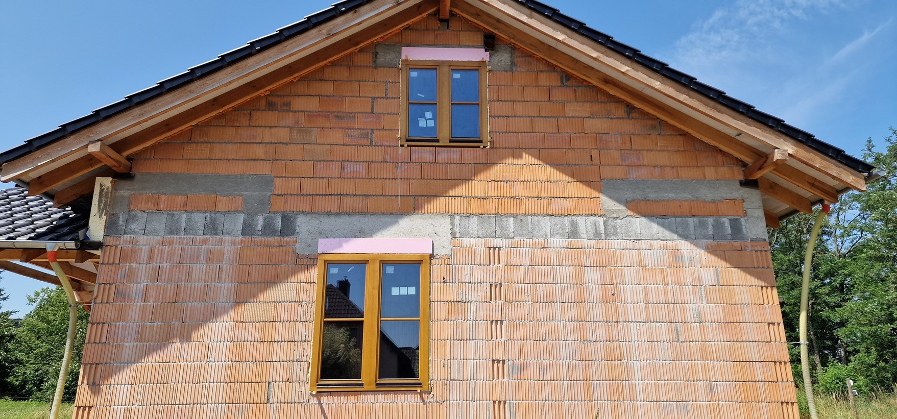 Surowy stan budynku z czerwonej cegły z zamontowanymi oknami z ramami w kolorze drewna, zabezpieczonymi różową pianką montażową, widoczne odbicie w szybach okiennych.