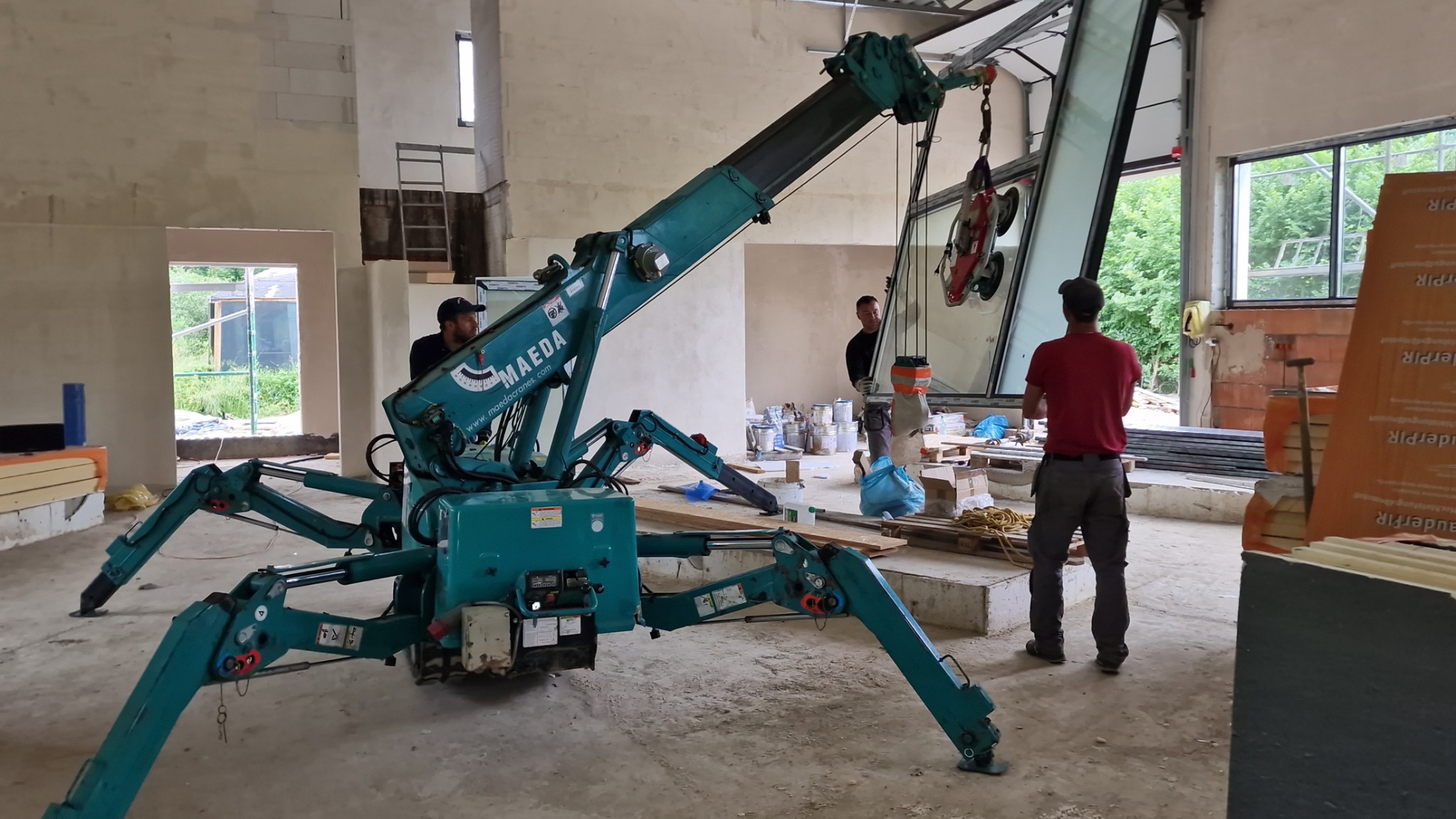 Montaż dużego okna z użyciem mini żurawia Maeda w surowym wnętrzu budynku; widoczni dwaj monterzy i operator żurawia.