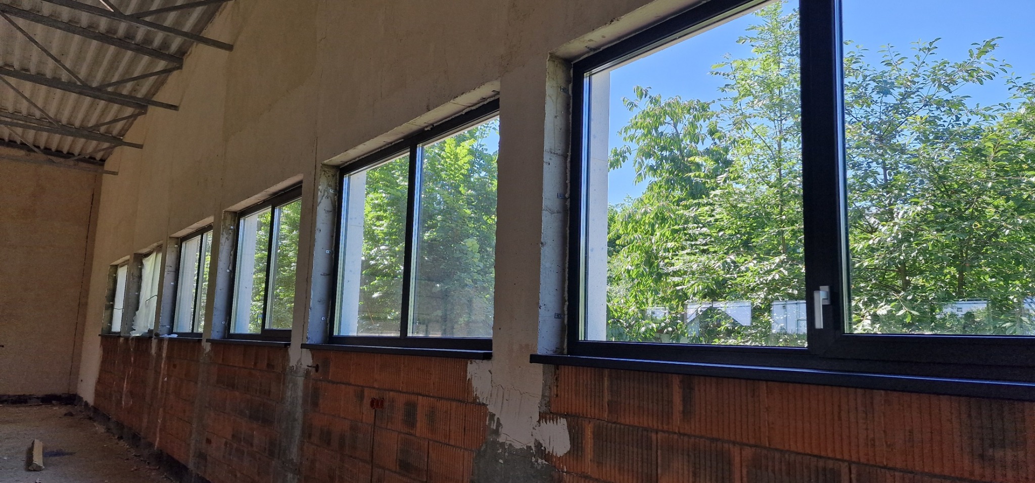 Rząd czarnych okien z widokiem na zieleń drzew, zamontowanych w surowym stanie budynku z ceglaną ścianą.