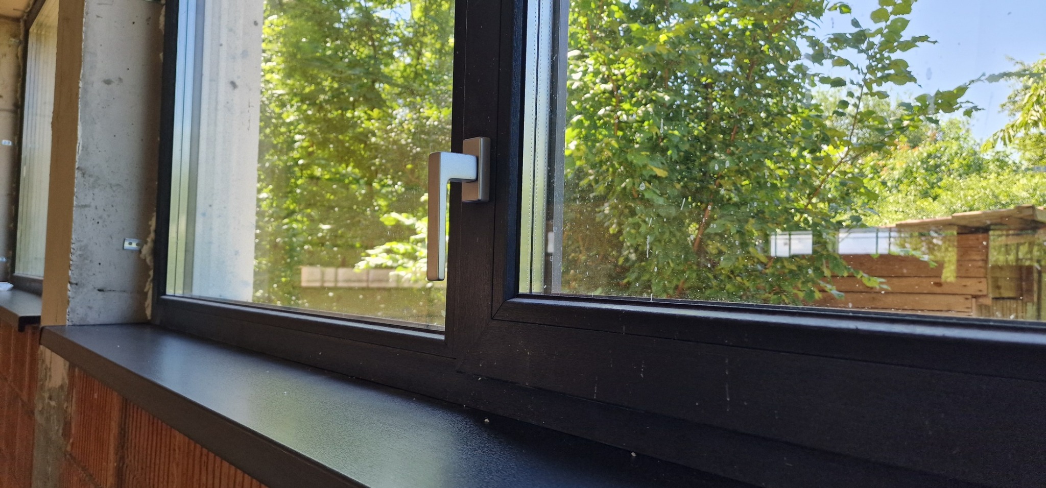 Ciemne okno PCV z widokiem na zielony ogród; surowy mur w tle podkreśla kontrast między nowoczesnym oknem a tradycyjnym budownictwem.