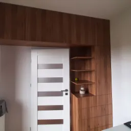 Zabudowa wnęki z szafkami, półkami i szufladami z drewna w ciepłym odcieniu, zintegrowana z białą, nowoczesną stolarką drzwiową z poziomymi przeszkleniami.