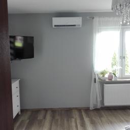 KOTECH Paweł Kot - Biały klimatyzator ścienny w szarej sypialni, widoczny telewizor na ścianie, biała komoda, okno z białymi firankami, parkiet na podłodze i ozdobny żyrandol