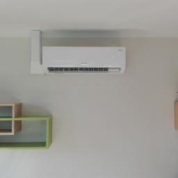 KOTECH Paweł Kot - Biały klimatyzator Rotenso zamontowany na ścianie w pokoju z minimalistycznym wystrojem, obok wiszące półki z dekoracjami.