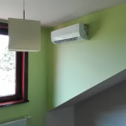 Biały klimatyzator zamontowany na zielonej ścianie w jasnym pomieszczeniu, widoczny grzejnik pod oknem i lampa wisząca.