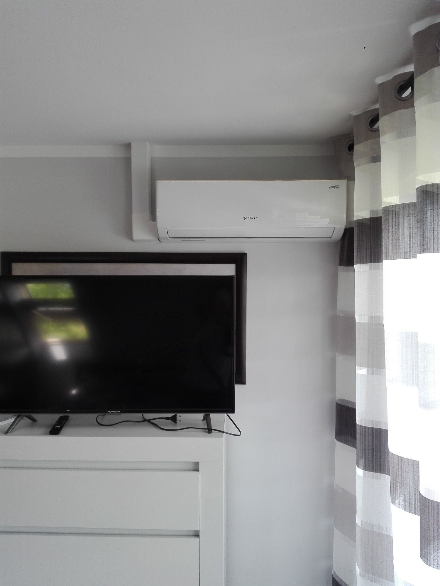 Biały klimatyzator Rotenso zamontowany nad telewizorem w nowoczesnym wnętrzu z szaro-białymi zasłonami.