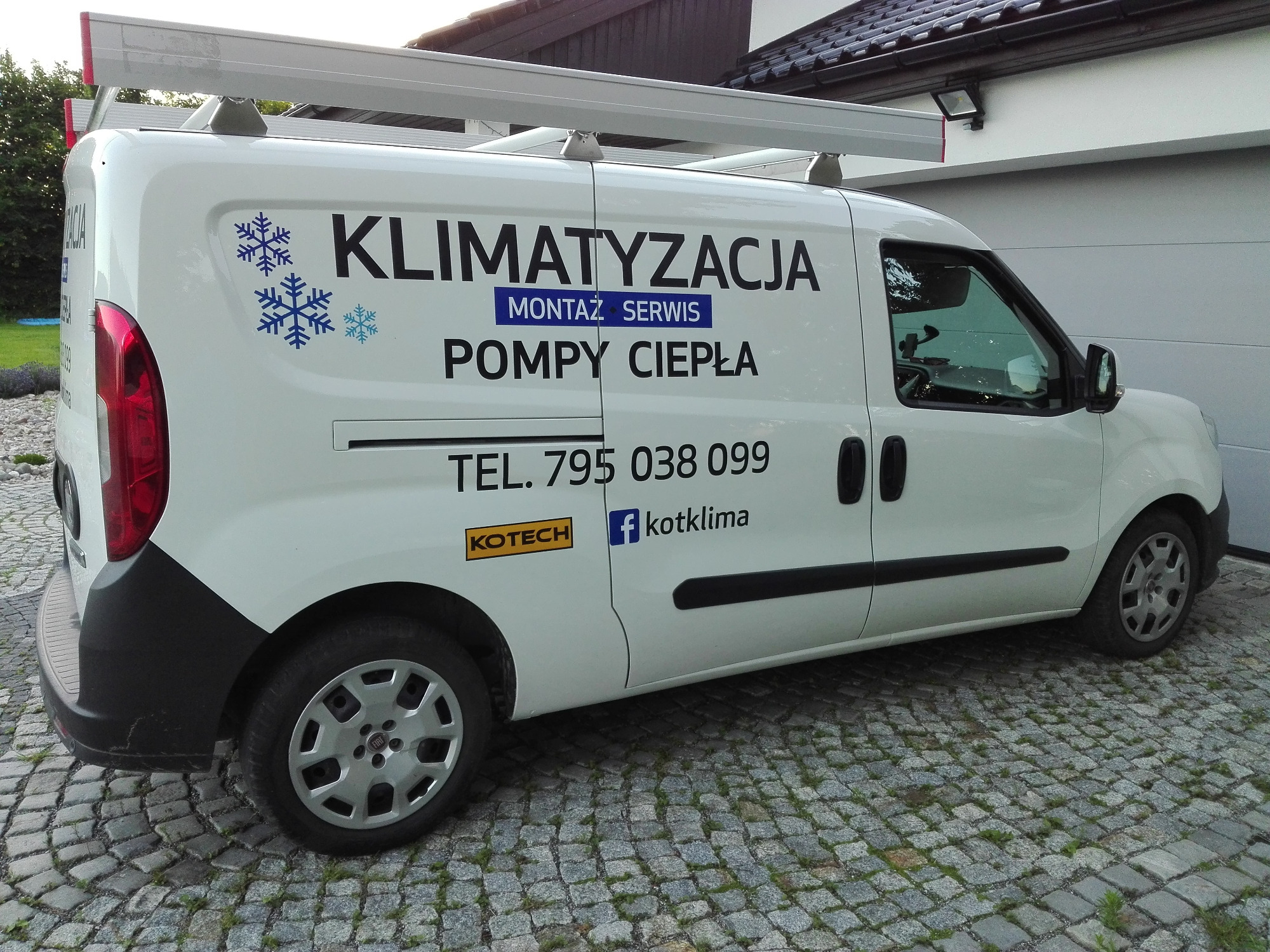 Biały samochód dostawczy z logo firmy świadczącej usługi montażu, serwisu klimatyzacji i pomp ciepła, zaparkowany na kostce brukowej przed garażem.