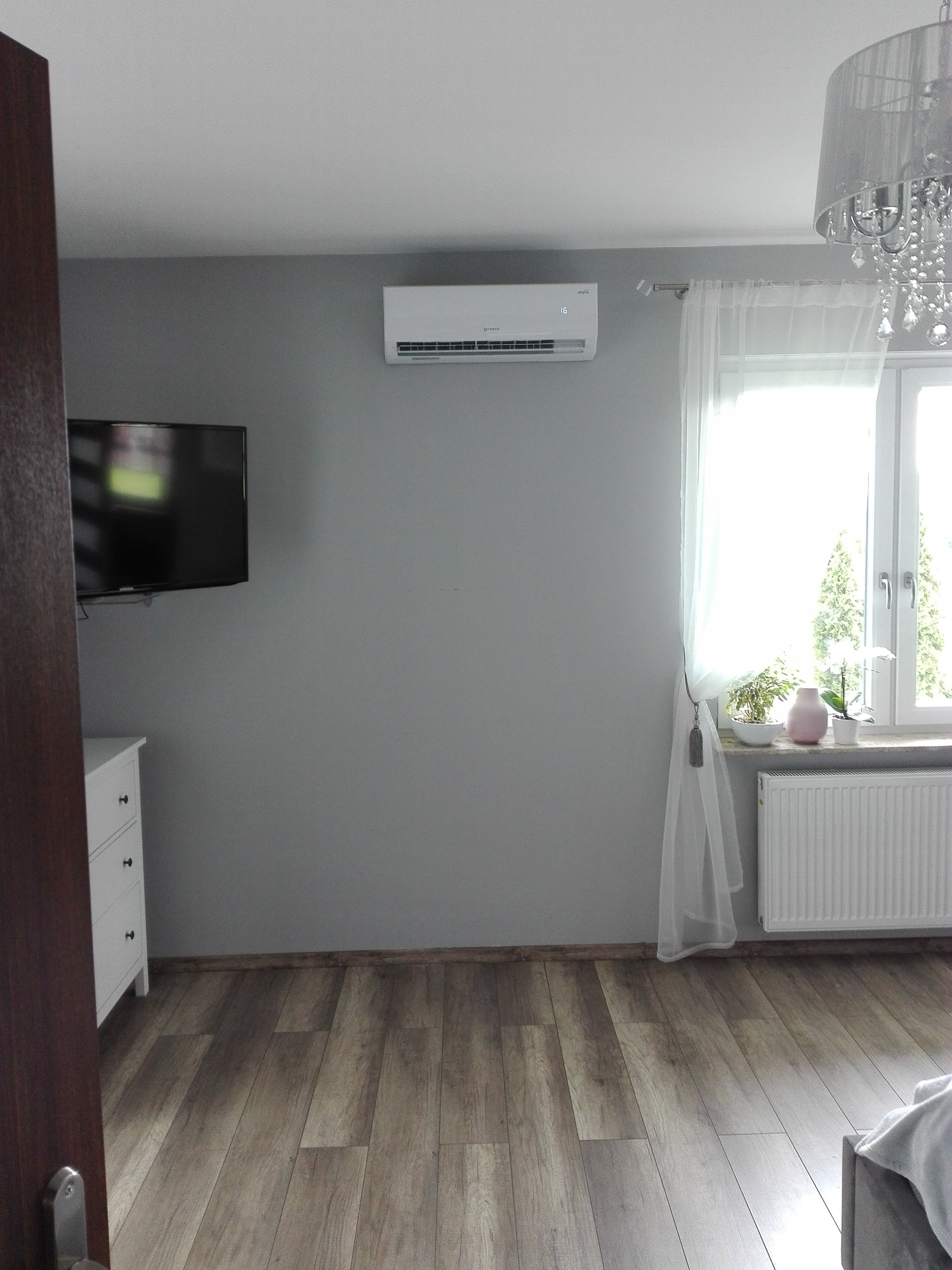 Biały klimatyzator ścienny w szarej sypialni, widoczny telewizor na ścianie, biała komoda, okno z białymi firankami, parkiet na podłodze i ozdobny żyrandol