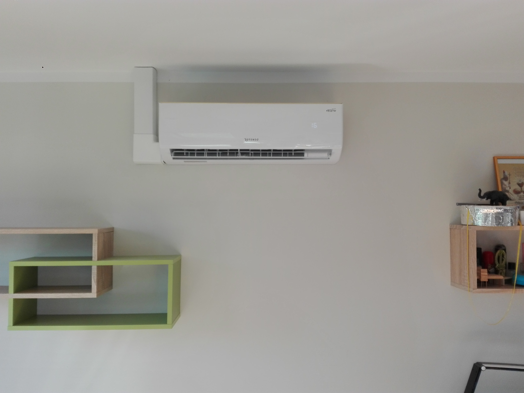 Biały klimatyzator Rotenso zamontowany na ścianie w pokoju z minimalistycznym wystrojem, obok wiszące półki z dekoracjami.