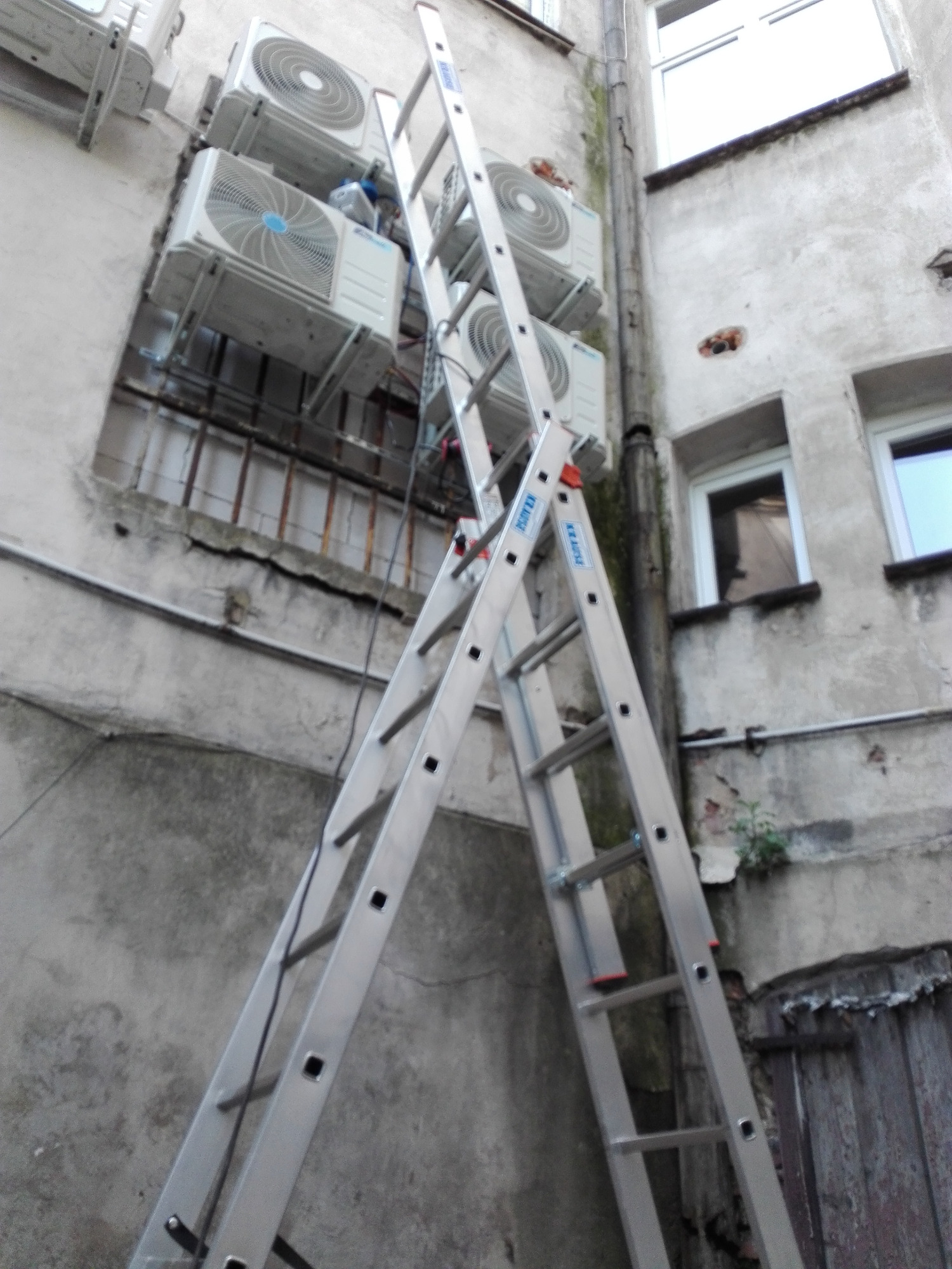 Zewnętrzne jednostki klimatyzacyjne zamontowane na elewacji budynku, widoczne drabiny aluminiowe ustawione do ściany, instalacja w trakcie montażu.