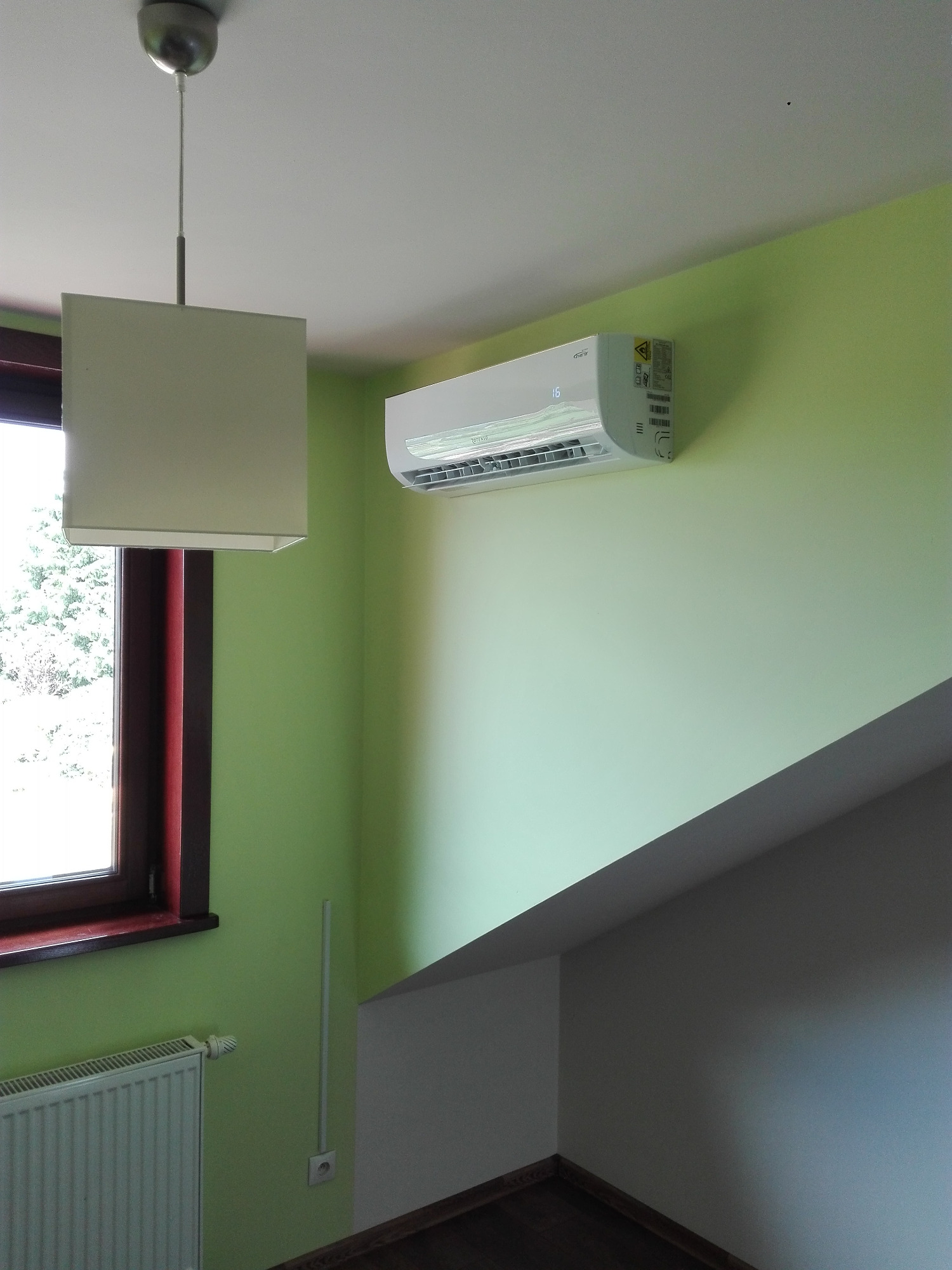Biały klimatyzator zamontowany na zielonej ścianie w jasnym pomieszczeniu, widoczny grzejnik pod oknem i lampa wisząca.