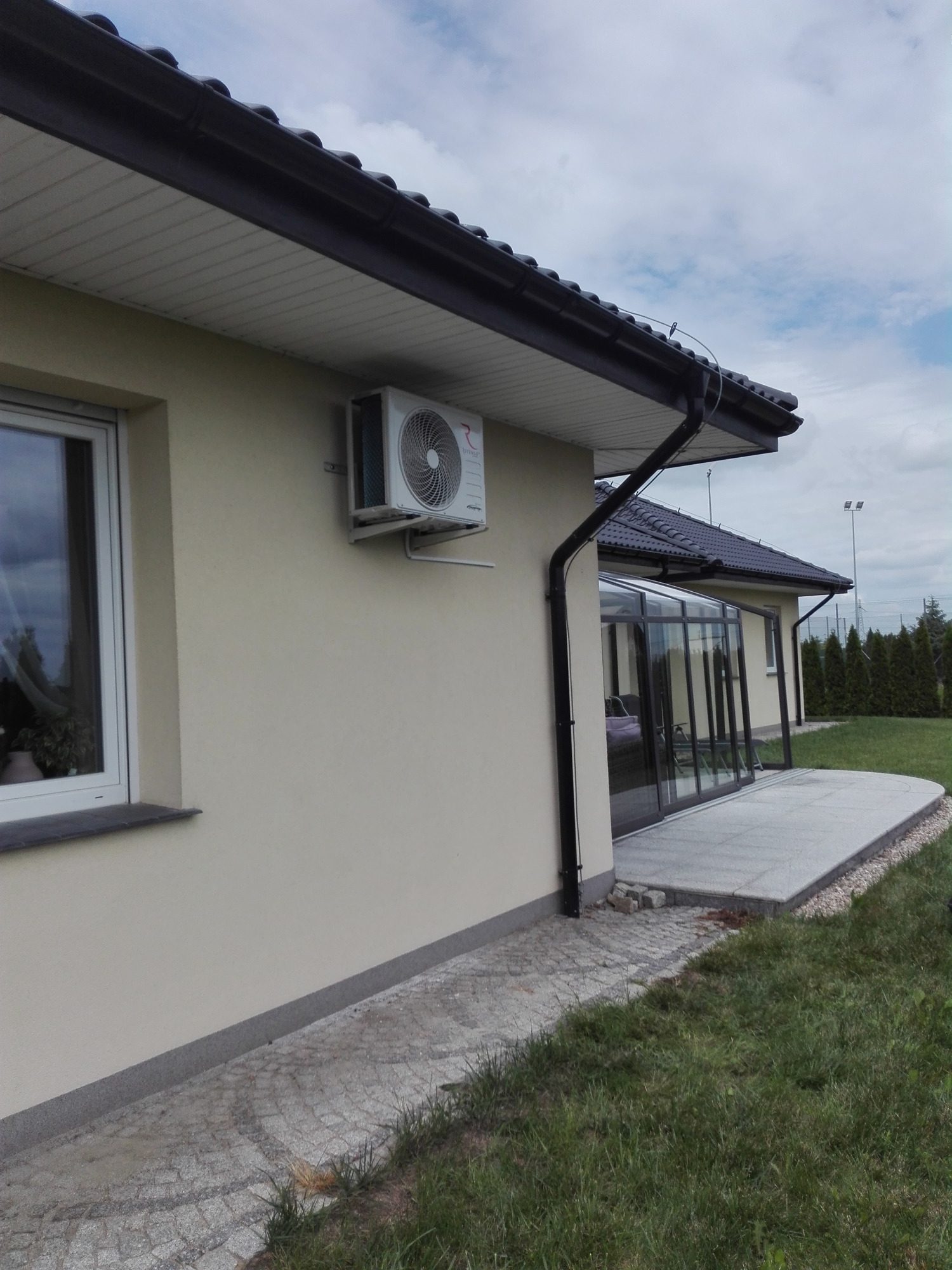 Biały klimatyzator zamontowany na elewacji domu jednorodzinnego z widoczną werandą i ogrodem w tle.