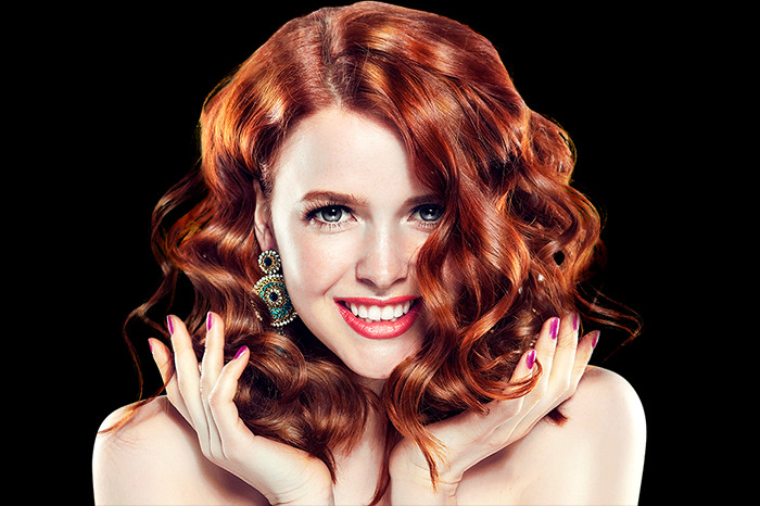 Portret uśmiechniętej kobiety z rudymi, kręconymi włosami, widoczne duże kolczyki z turkusowymi akcentami i fioletowy manicure na tle czerni.