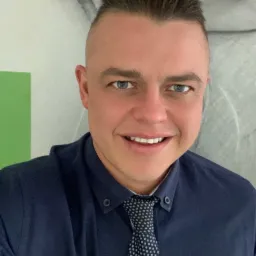 Uśmiechnięty mężczyzna w granatowej koszuli i krawacie w kropki, z krótkimi włosami i niebieskimi oczami, pozujący na tle abstrakcyjnej grafiki ściennej i zielonego elementu dekoracyjnego.