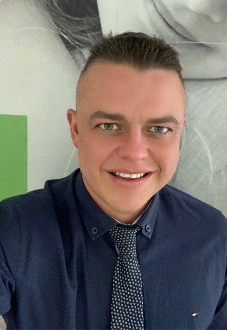 Uśmiechnięty mężczyzna w granatowej koszuli i krawacie w kropki, z krótkimi włosami i niebieskimi oczami, pozujący na tle abstrakcyjnej grafiki ściennej i zielonego elementu dekoracyjnego.