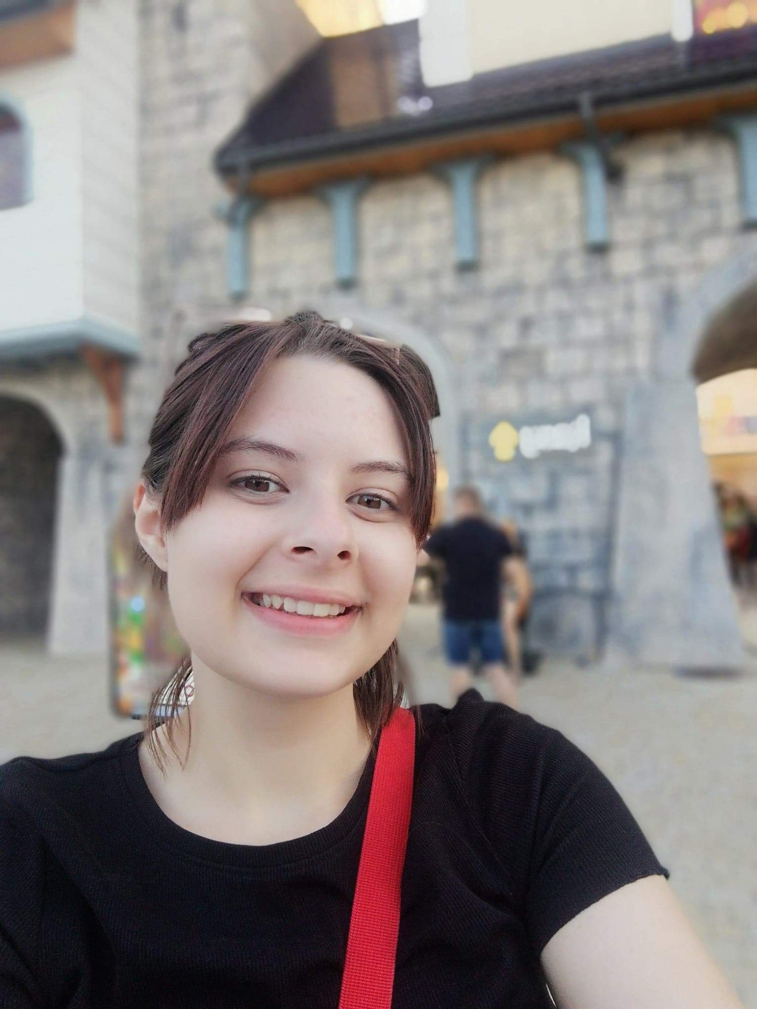 Uśmiechnięta młoda kobieta w czarnej bluzce z czerwoną torbą na ramieniu, na tle budynku stylizowanego na zamek z kamienia.