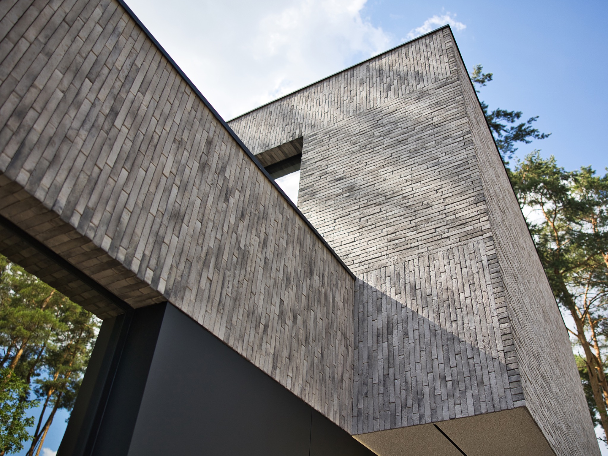 Nowoczesna elewacja domu z szarej cegły klinkierowej, widok pod kątem z dołu, z fragmentem niebieskiego nieba i zieleni w tle.