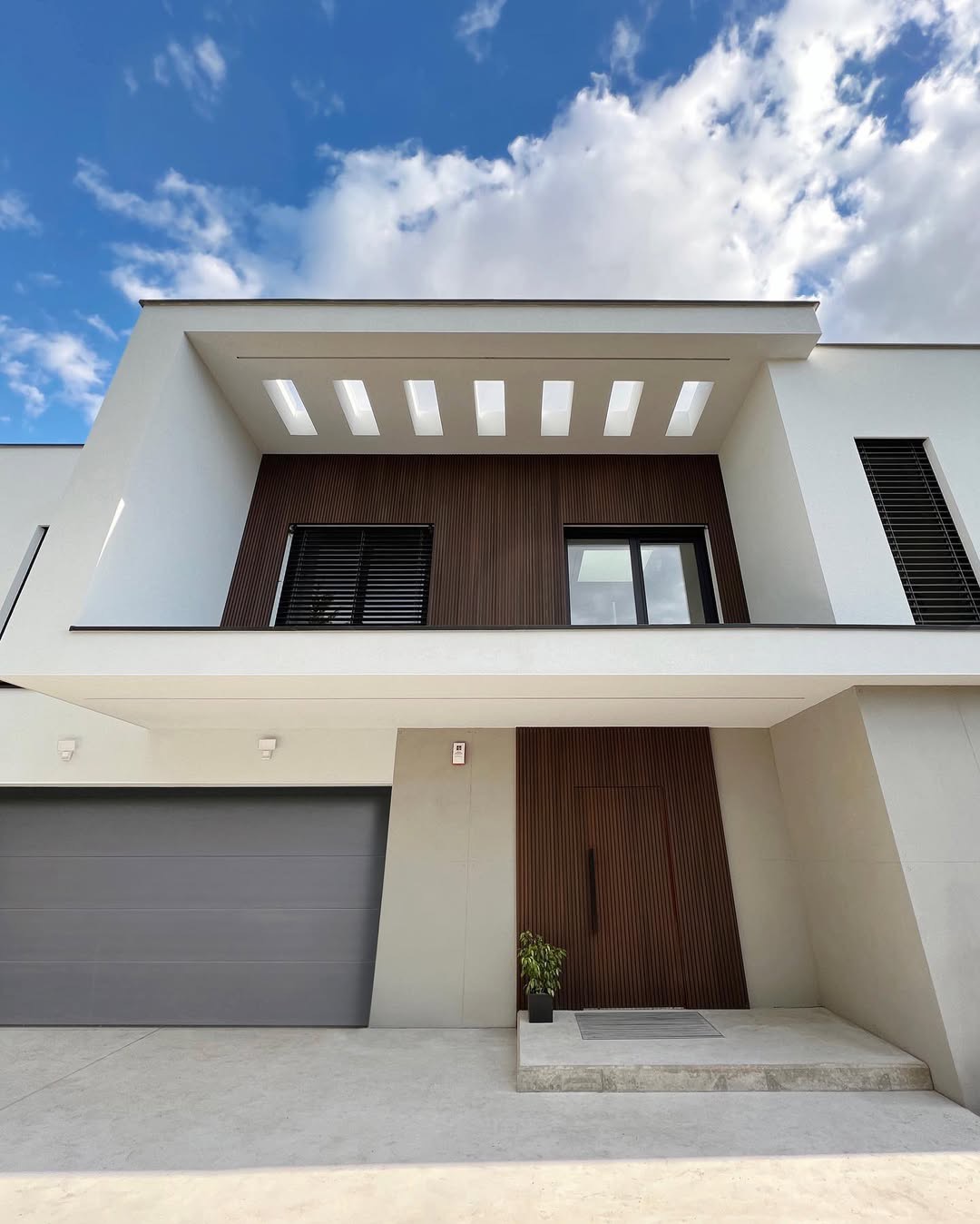 Nowoczesny dom jednorodzinny z białą elewacją, drewnianymi akcentami i szarą bramą garażową, sfotografowany w słoneczny dzień z perspektywy żabiej.