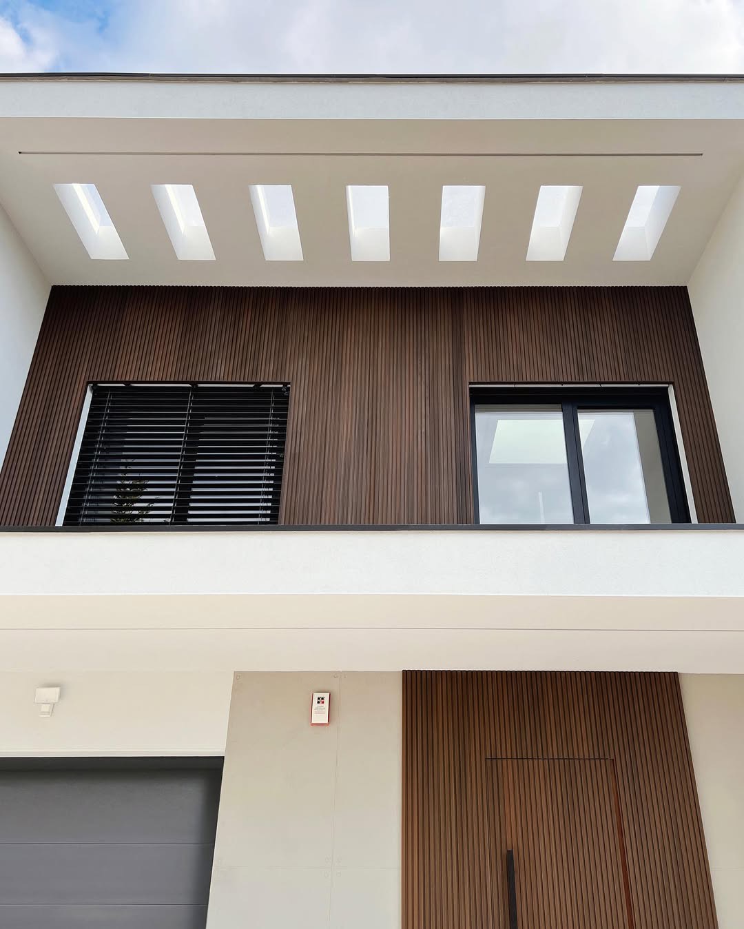 Nowoczesna elewacja domu z drewnianymi panelami, oknami z żaluzjami i oświetleniem w suficie podwieszanym.