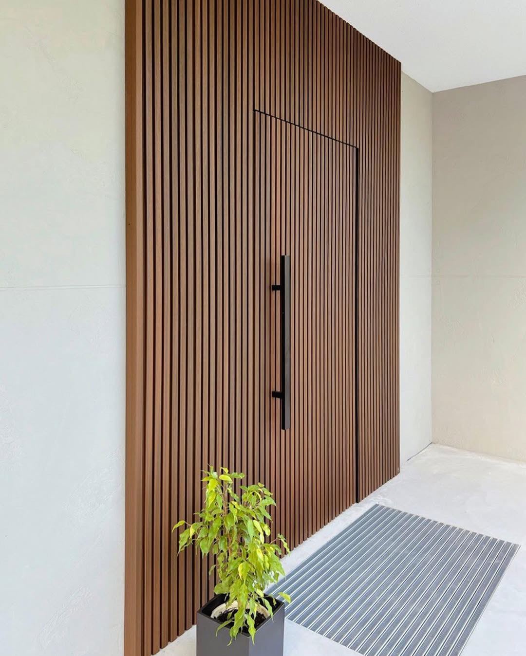 Nowoczesna elewacja z drewnianych lameli z ukrytymi drzwiami wejściowymi i minimalistycznym uchwytem, uzupełniona rośliną doniczkową w kwadratowej donicy i metalową wycieraczką.