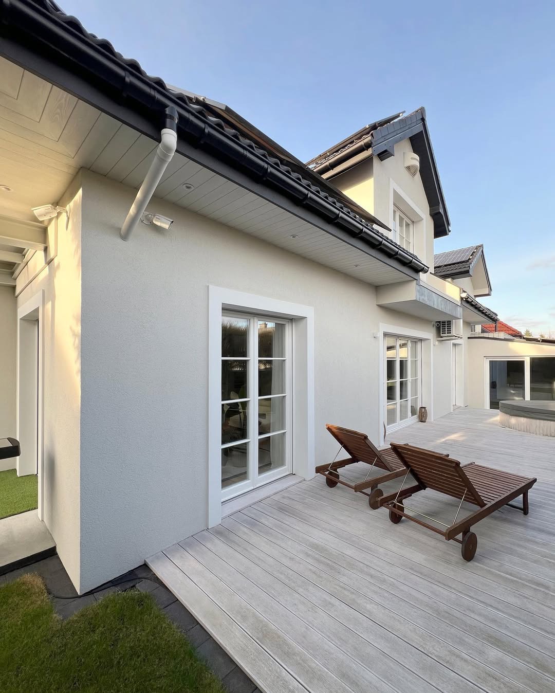 Nowoczesny dom z jasną elewacją, drewnianym tarasem i dwoma leżakami, widok z boku w słoneczny dzień.