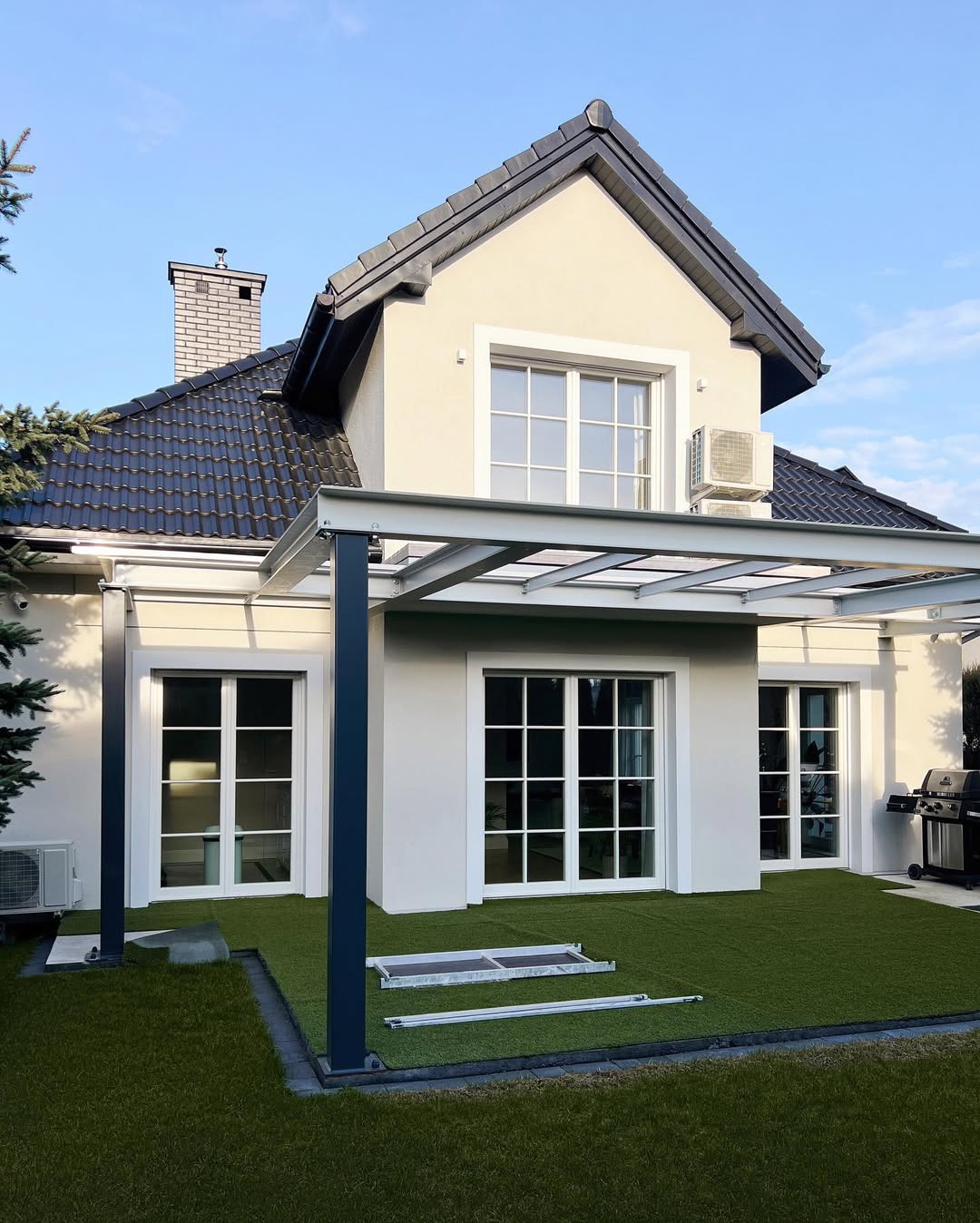 Jasna elewacja domu jednorodzinnego z ciemną dachówką, widoczne okna z białymi ramami, klimatyzator na ścianie, zadaszenie tarasu z metalową konstrukcją i sztuczna trawa na patio, fragment komina...