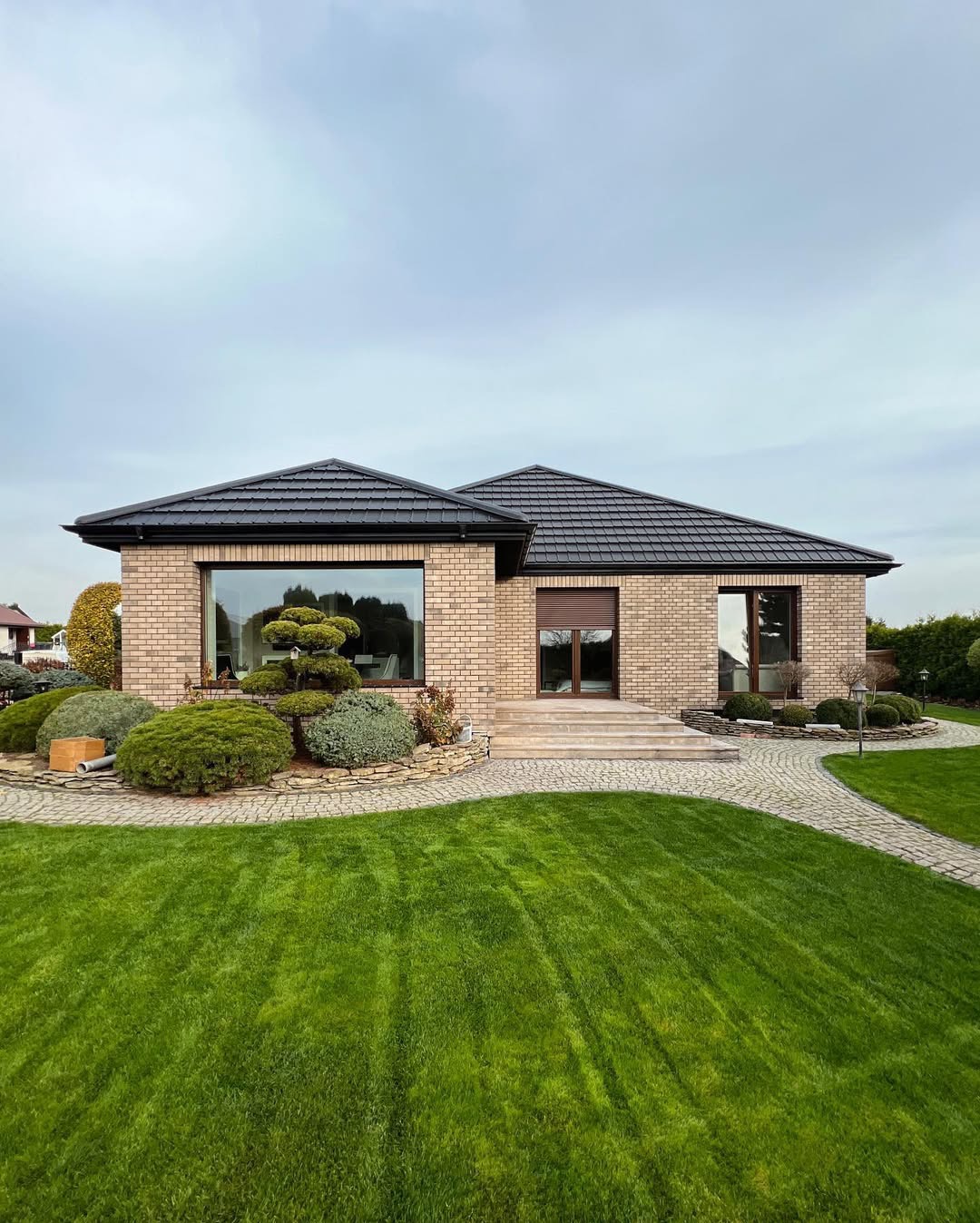 Parterowy dom z cegły klinkierowej, czarną dachówką i dużym oknem panoramicznym, otoczony zadbanym trawnikiem i ozdobnymi krzewami.