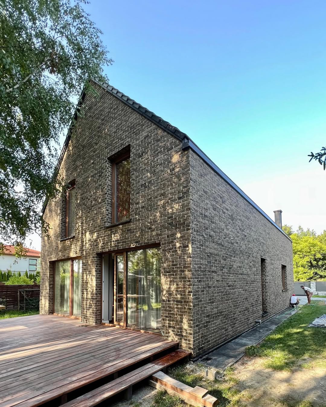 Nowoczesny dom z elewacją z ciemnej cegły klinkierowej, widoczny taras z desek i fragment ogrodu, dwóch pracowników przy elewacji z tyłu budynku.