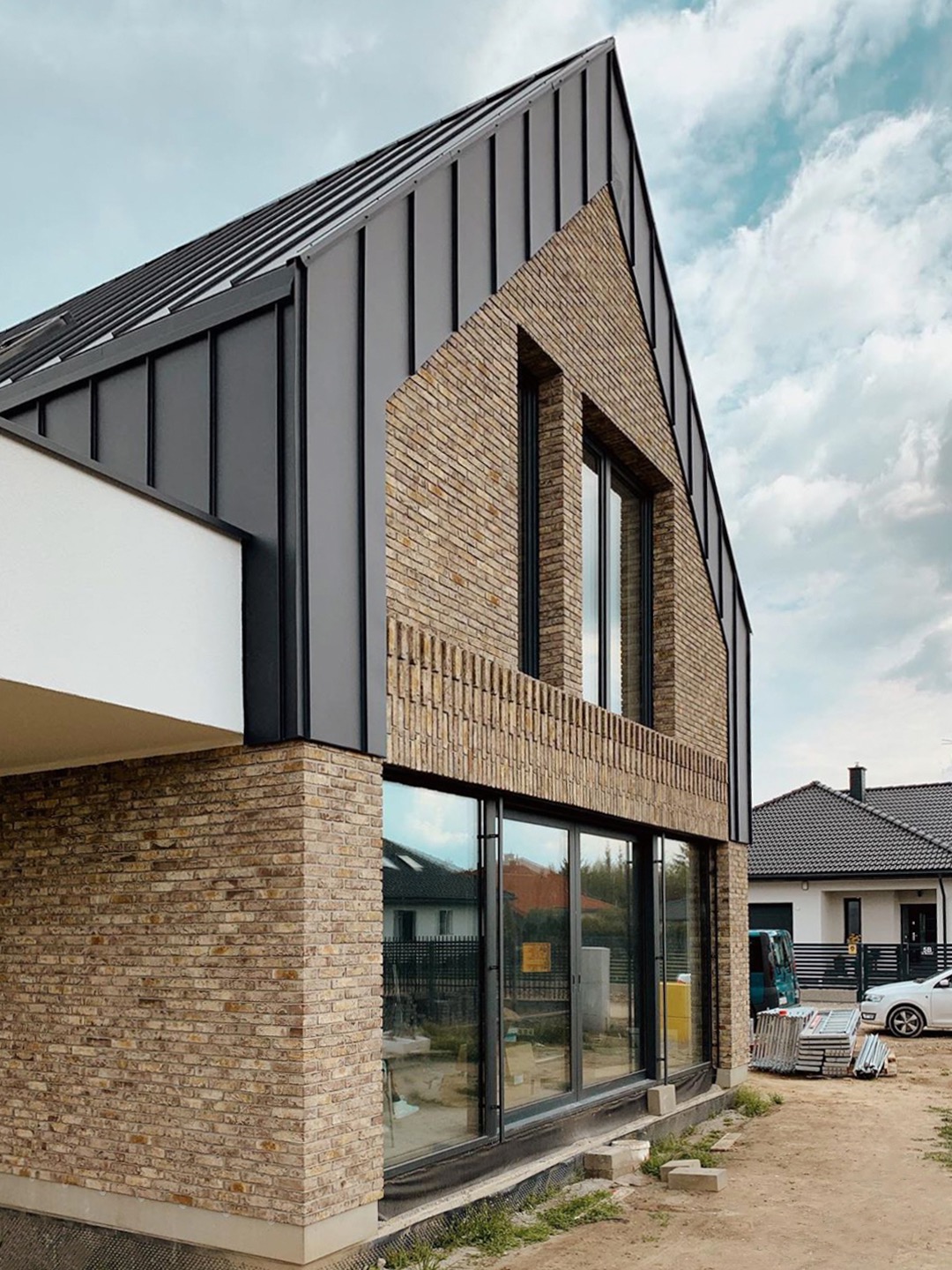 Nowoczesny dom jednorodzinny z elewacją z cegły klinkierowej i grafitowym dachem z blachy na rąbek stojący, duże okna z czarnymi ramami, widoczne przygotowania do zagospodarowania terenu.