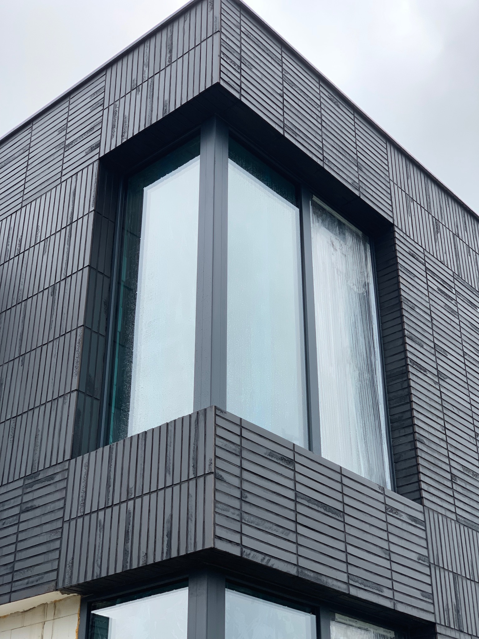 Nowoczesna elewacja budynku wykończona ciemnymi, wąskimi płytkami ceramicznymi, z dużym narożnym oknem, widziana pod kątem od dołu.
