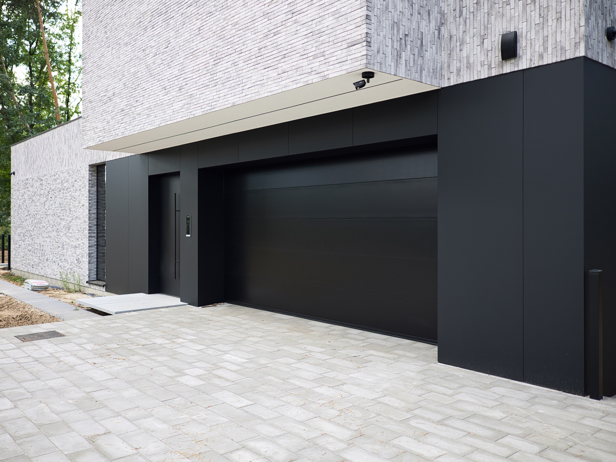 Nowoczesny dom z elewacją z białej cegły, czarną bramą garażową segmentową i minimalistycznym oświetleniem zewnętrznym.