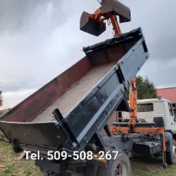 Czarno-pomarańczowa ciężarówka wywrotka z podniesioną skrzynią ładunkową i ramieniem z łyżką, stojąca na trawiastym terenie pod pochmurnym niebem. Na skrzyni widoczne ślady użytkowania.