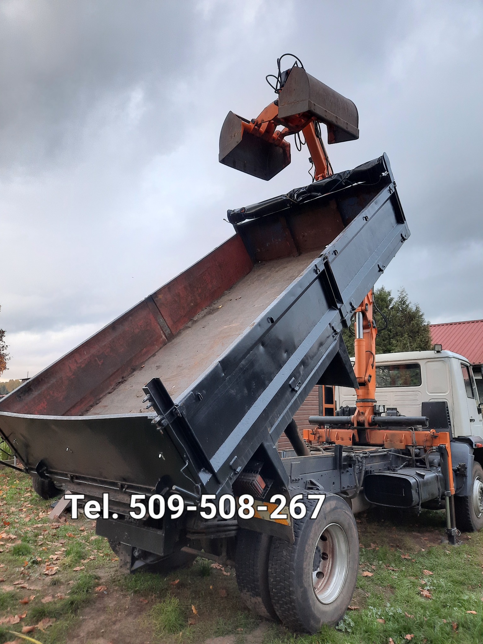 Czarno-pomarańczowa ciężarówka wywrotka z podniesioną skrzynią ładunkową i ramieniem z łyżką, stojąca na trawiastym terenie pod pochmurnym niebem. Na skrzyni widoczne ślady użytkowania.