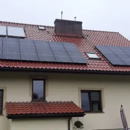 Dom jednorodzinny z panelami słonecznymi na czerwonym dachu w pochmurny dzień; widoczne trzy zestawy paneli w różnych rozmiarach.