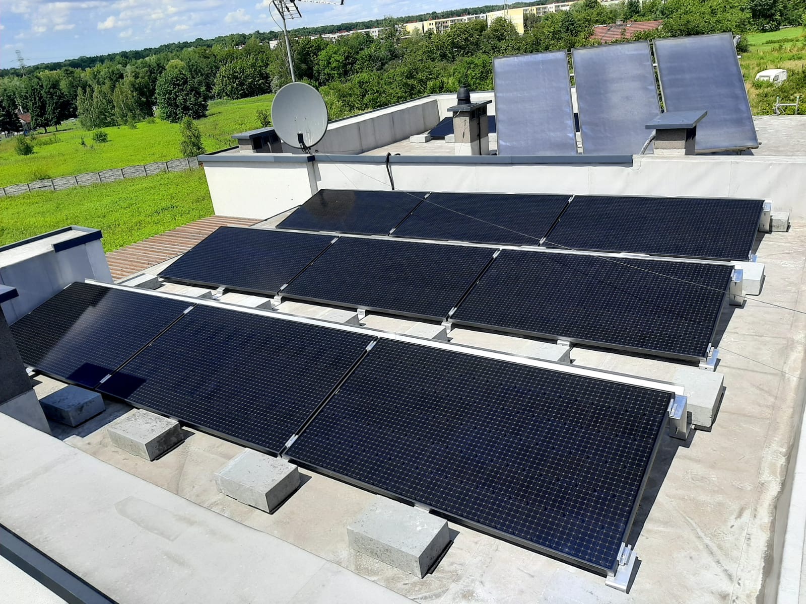 Instalacja fotowoltaiczna na płaskim dachu z widocznymi betonowymi elementami podtrzymującymi panele, antena satelitarna w tle.