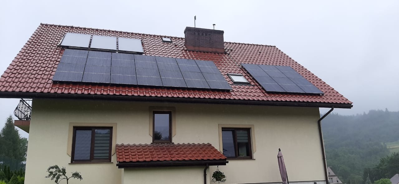 Dom jednorodzinny z panelami słonecznymi na czerwonym dachu w pochmurny dzień; widoczne trzy zestawy paneli w różnych rozmiarach.