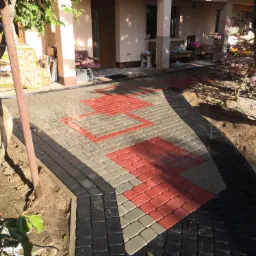 Ułożona ścieżka z kostki brukowej w trzech kolorach: czarnym, szarym i czerwonym, prowadząca do wejścia domu z widocznym fragmentem ogrodu i drzewa.