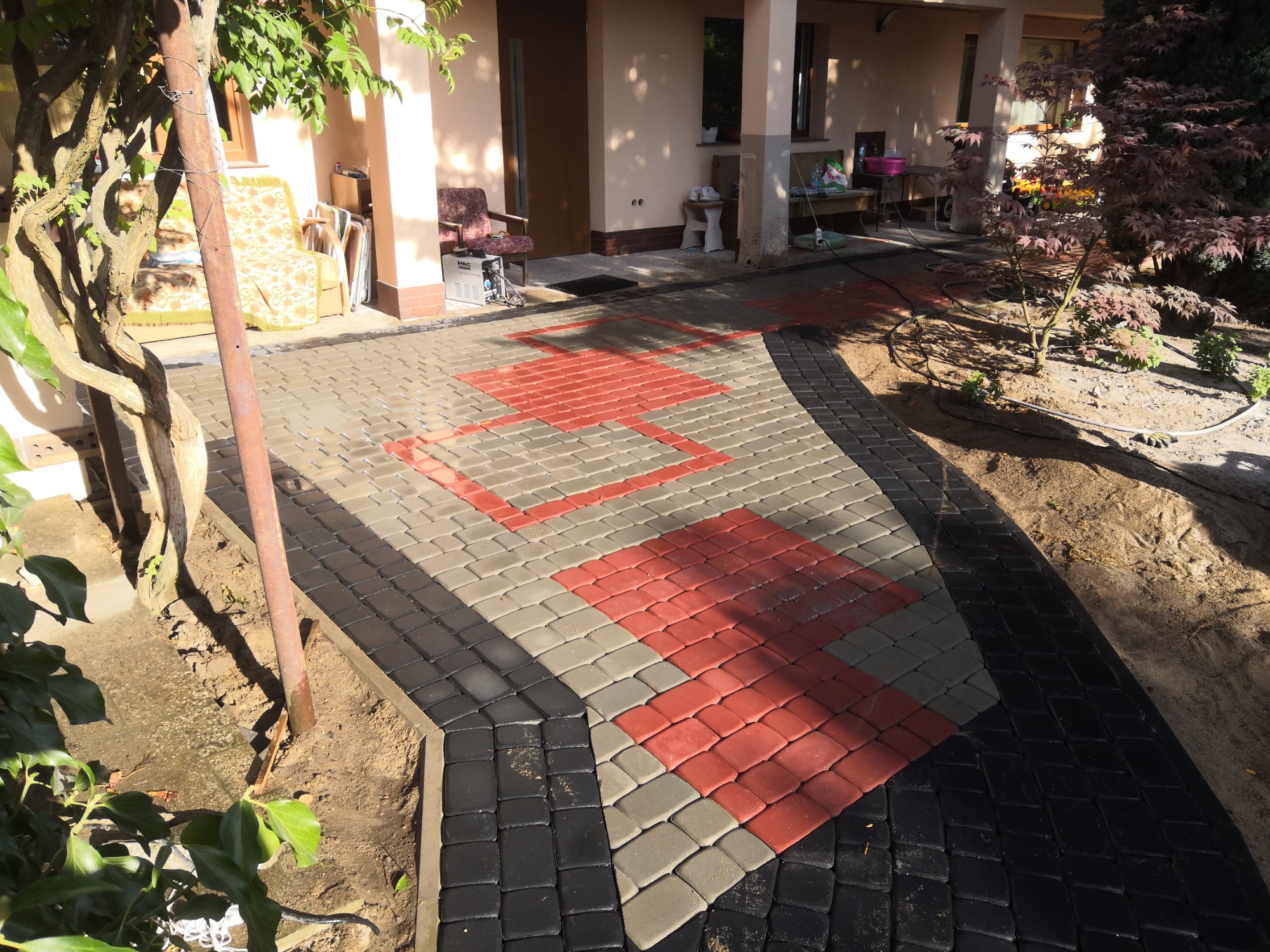 Ułożona ścieżka z kostki brukowej w trzech kolorach: czarnym, szarym i czerwonym, prowadząca do wejścia domu z widocznym fragmentem ogrodu i drzewa.