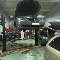 Dako Auto - Szary samochód na podnośniku w warsztacie; otwarte maski dwóch aut, widoczne narzędzia i części zamienne na podłodze w kratkę.