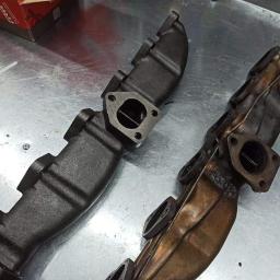 Dako Auto - Dwa kolektory wydechowe: nowy, pomalowany na czarno oraz używany, z widocznym nagarem i śladami spawania, leżące na metalowym stole warsztatowym.