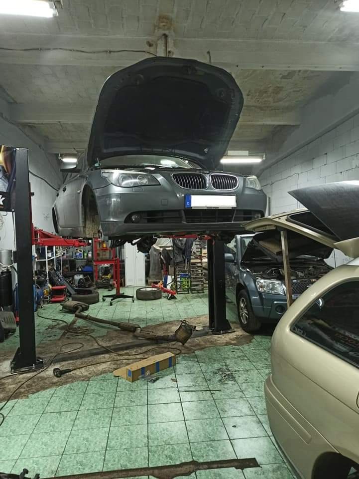 Szary samochód na podnośniku w warsztacie; otwarte maski dwóch aut, widoczne narzędzia i części zamienne na podłodze w kratkę.