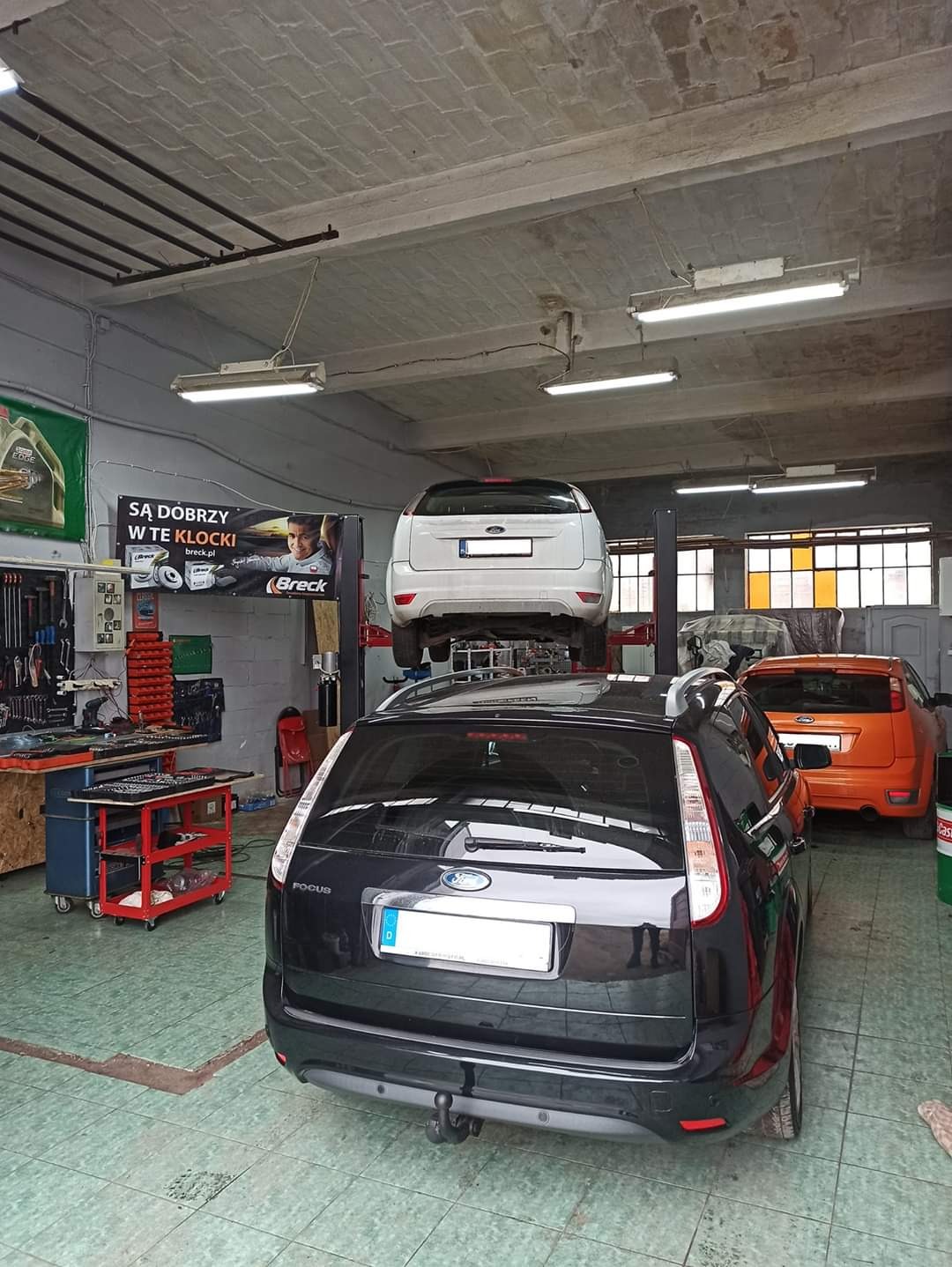 Wnętrze warsztatu samochodowego z dwoma Fordami Focus: czarnym kombi na pierwszym planie i białym hatchbackiem na podnośniku. W tle widoczny pomarańczowy samochód i narzędzia warsztatowe.