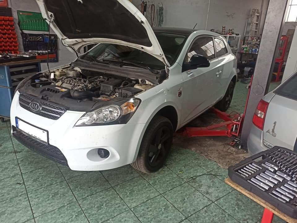 Biała Kia Ceed na podnośniku w warsztacie samochodowym z otwartą maską, widoczne narzędzia i wyposażenie warsztatu.