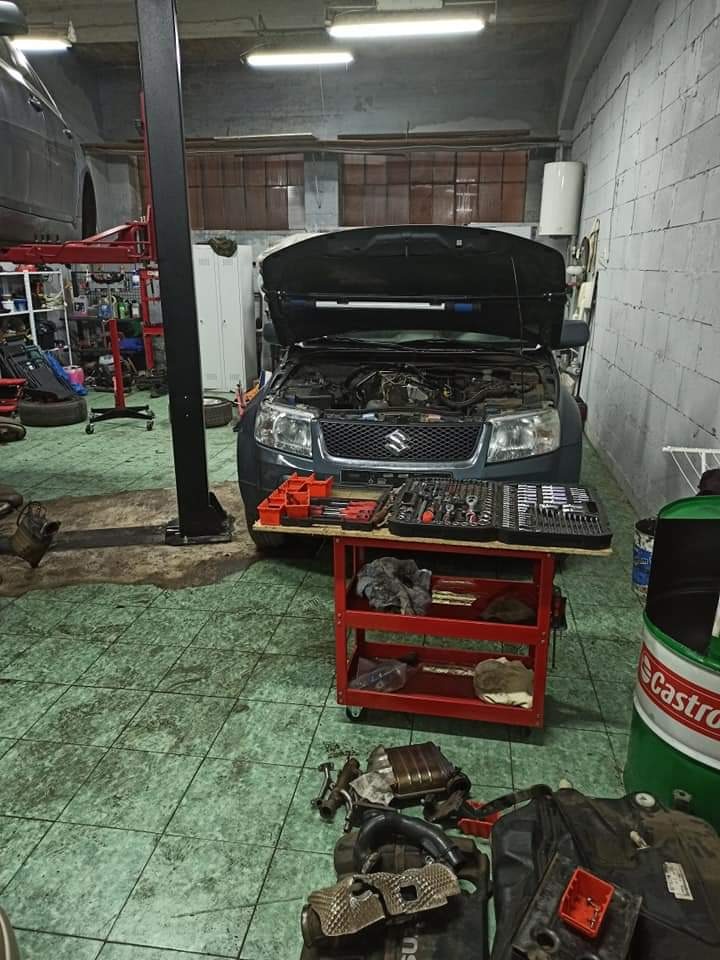 Szary samochód Suzuki z otwartą maską na stanowisku warsztatowym, narzędzia na wózku obok, widoczna puszka Castrol i podnośnik samochodowy w tle.