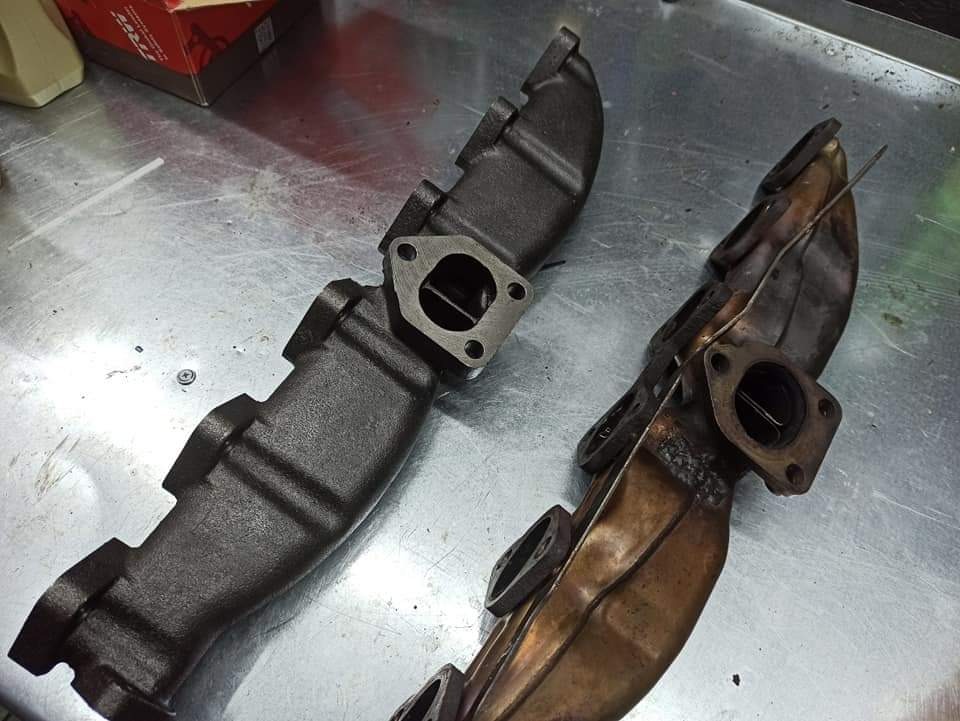 Dwa kolektory wydechowe: nowy, pomalowany na czarno oraz używany, z widocznym nagarem i śladami spawania, leżące na metalowym stole warsztatowym.