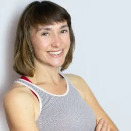 Uśmiechnięta kobieta w sportowej koszulce z rękami skrzyżowanymi na tle białej ściany, prezentująca zdrowe zęby.